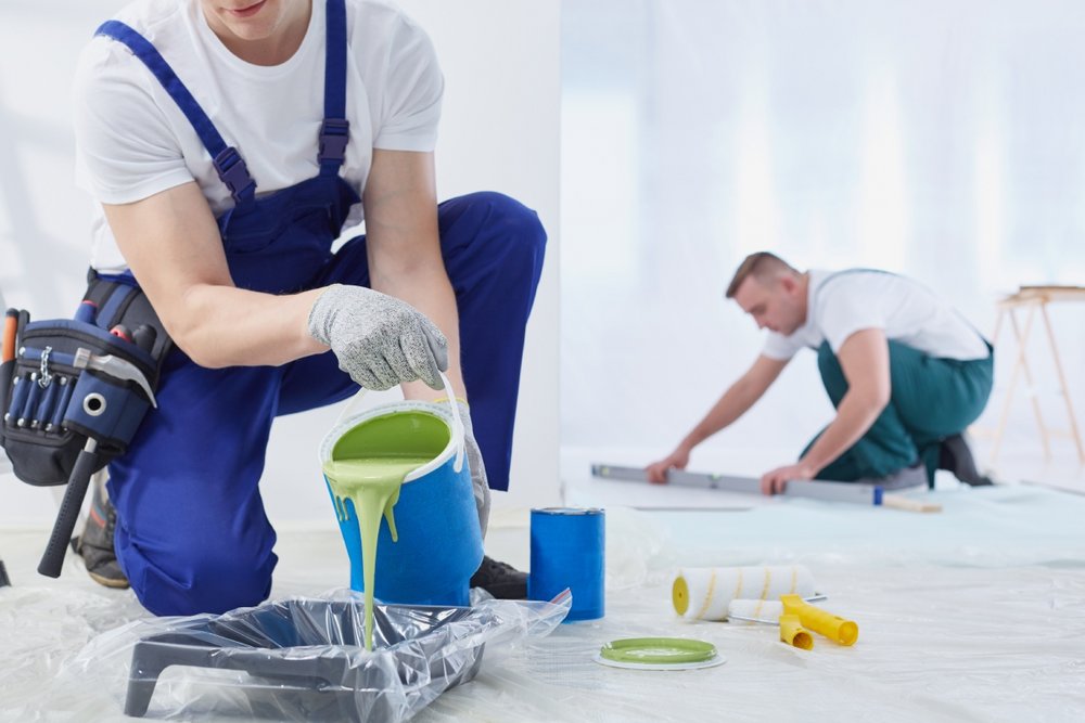 Arbeiter im Blaumann gießt Farbe um, Kollege im Hintergrund benutzt eine Wasserwaage